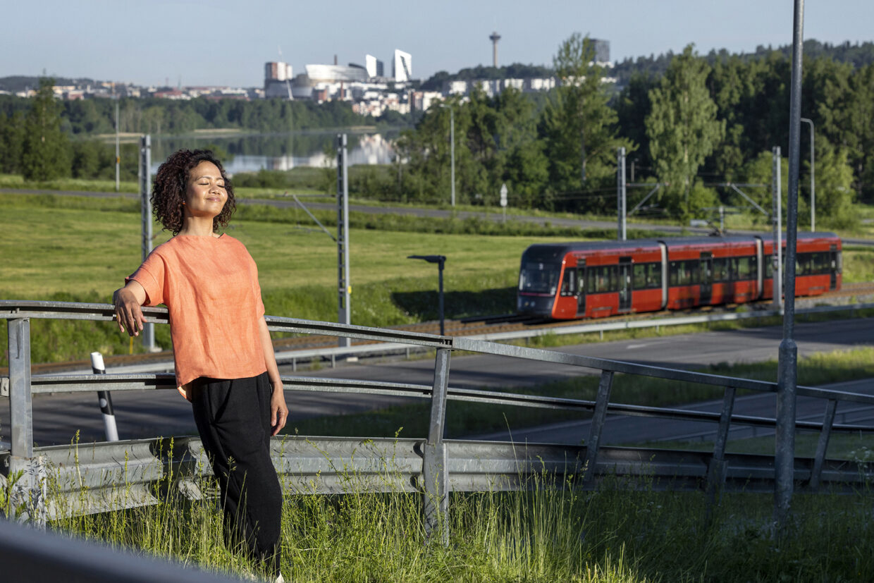Nainen nojaa kaiteeseen ja nauttii auringonpaisteesta. Taustalla näkyy punainen ratikka.